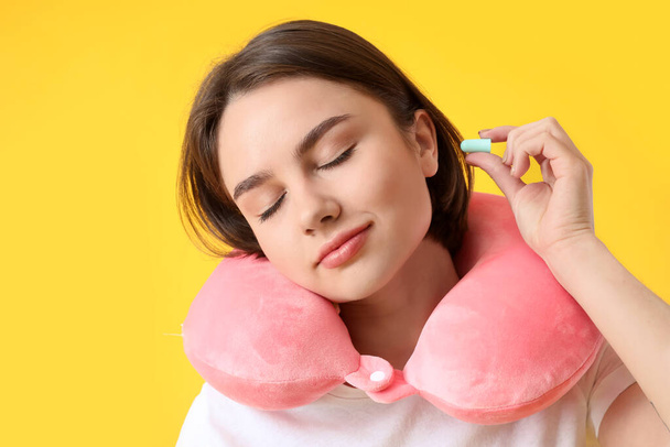 Young woman with neck pillow putting ear plug on yellow background - Photo, Image