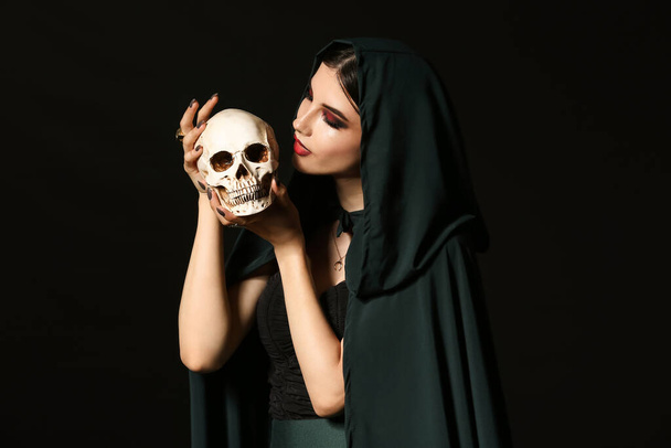 Young witch with human skull on dark background - Φωτογραφία, εικόνα