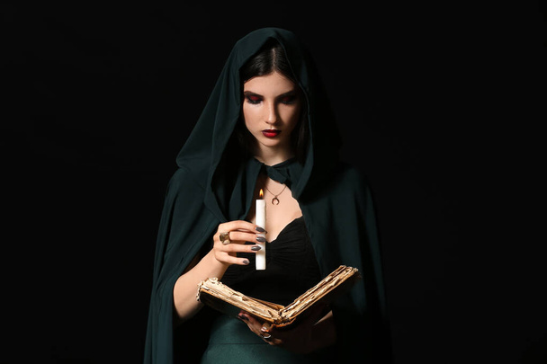 Young witch with spell book and candle on dark background - Фото, изображение