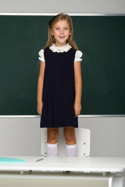 Smiling blonde female student in school uniform stands on a chair in front of the blackboard. School and education concept - Фото, изображение