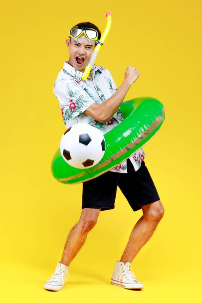 Jeune homme asiatique attrayant en chemise hawaïenne blanche portant un masque de plongée jaune et un anneau de natation vert autour de sa taille tenant le ballon sur fond jaune. Concept pour des vacances à la plage. - Photo, image