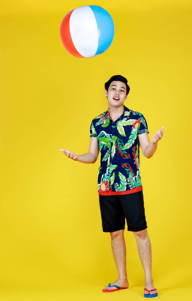 Jeune homme asiatique attrayant en chemise hawaïenne bleue et verte jetant balle de plage à l'air sur fond jaune. Concept pour des vacances à la plage. - Photo, image