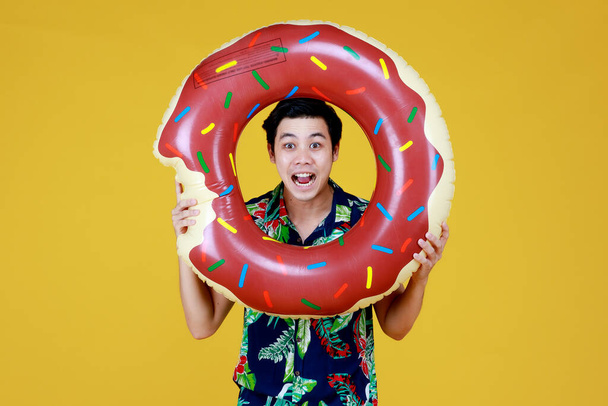 Young attractive Asian man wearing green and blue Hawaiian shirt with donut swim ring infront his face going for beach vacation against yellow background. - Photo, Image