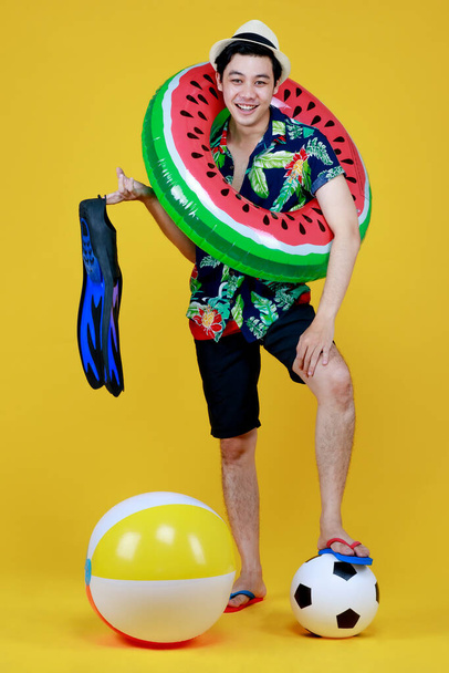 Jeune homme asiatique attrayant portant une chemise hawaïenne verte et bleue et un anneau de natation de pastèque autour de son corps tenant une nageoire bleue et une balle de plage allant pour des vacances à la plage sur fond jaune. - Photo, image