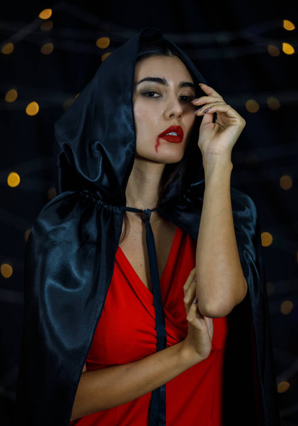 Νεαρή μυστηριώδης γυναίκα με αίμα στα κόκκινα χείλη φορώντας μαύρο μανδύα με κουκούλα αγγίζοντας το πηγούνι και κοιτάζοντας την κάμερα - Φωτογραφία, εικόνα