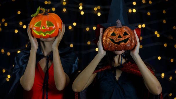 Unrecognizable girlfriends in witch costumes covering faces with pumpkin heads during night Halloween party - 写真・画像