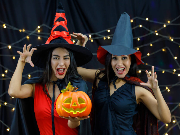 Jonge vrouwen in heksenhoeden en -mantels met pompoenkop die naar de camera kijken en enge gezichten en boe-gebaren doen terwijl ze samen Halloween vieren - Foto, afbeelding