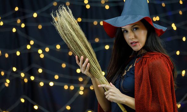 Vista lateral da encantadora jovem senhora em chapéu de bruxa e vassoura de equitação de capa e olhando para a câmera durante a celebração de Halloween da noite - Foto, Imagem