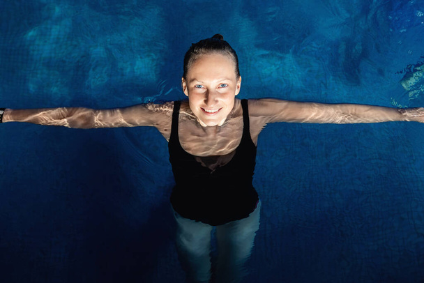 Над верхним видом портрет молодой взрослой кавказской модели в спортивном черном купальнике стоит чистая голубая вода крытого бассейна в темное вечернее время. Расслабьтесь гармония и оздоровительная терапия - Фото, изображение
