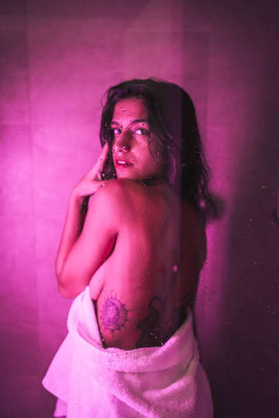 Lifestyle, sexy brunette Caucasian girl with towel at the end of the shower with a sensual look, with pink led light, portraits inside the shower from the back - Photo, Image