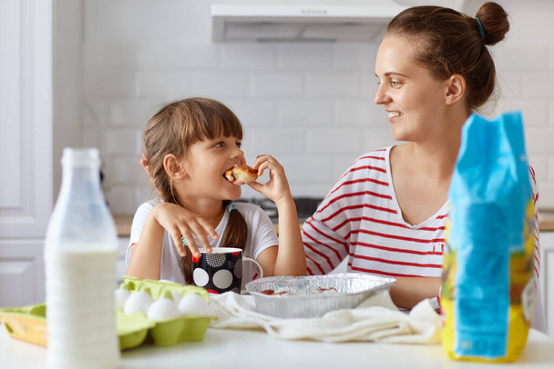 Portrait of young woman and her little cute daughter sitting at table and eating cakes and cookies on kitchen and drinking beverage, having fun together while enjoying freshly baked pastries. - Photo, Image