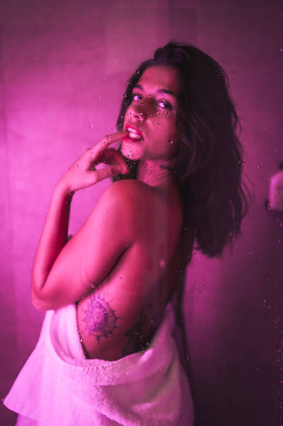 Lifestyle, sexy brunette Caucasian girl with towel at the end of the shower with sensual gaze, with pink led light, portraits inside the shower with bare back - Φωτογραφία, εικόνα