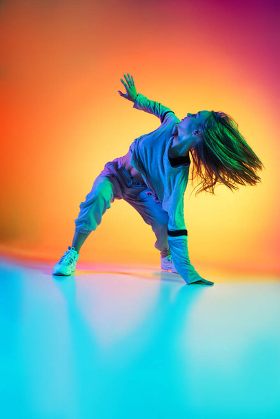 Sportive flexible girl dancing hip-hop in stylish clothes on colorful background at dance hall in neon light. Youth culture, movement, style and fashion, action. - Valokuva, kuva