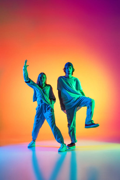 Young dancers, stylish emotive girl and boy dancing hip-hop in casual sports youth clothes on gradient multi colored background at dance hall in neon light. - Fotografie, Obrázek