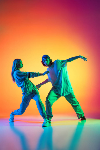 Young hip-hop dancers, stylish emotive girl and boy in action and motion in casual sports youth clothes on gradient multi colored background at dance hall in neon light. - Foto, Imagen