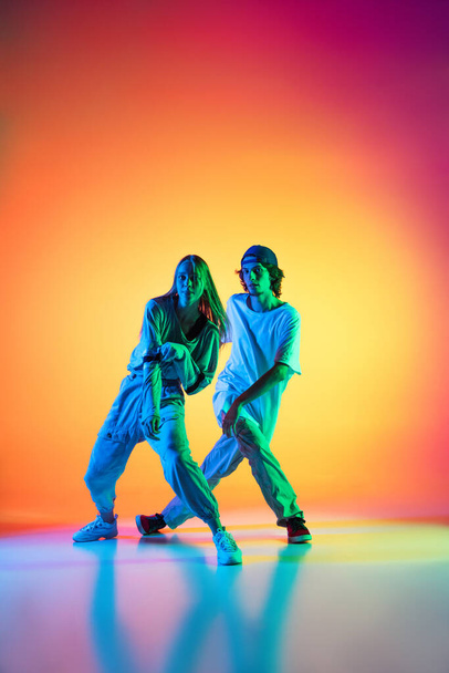 Young hip-hop dancers, stylish emotive girl and boy in action and motion in casual sports youth clothes on gradient multi colored background at dance hall in neon light. - Фото, изображение