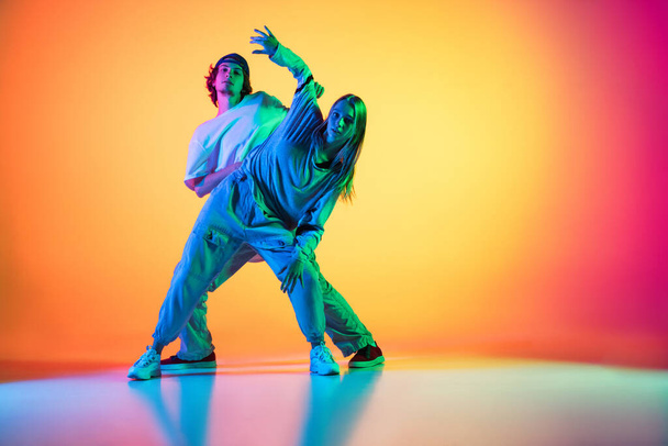 Young people, hip-hop dancers, stylish emotive girl and boy in action and motion in casual sports youth clothes on gradient multi colored background at dance hall in neon light. - Fotografie, Obrázek