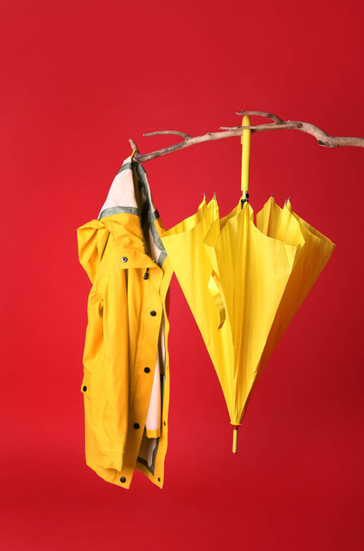 Geschlossener gelber Regenschirm und stylischer Regenmantel hängen am Ast vor rotem Hintergrund - Foto, Bild