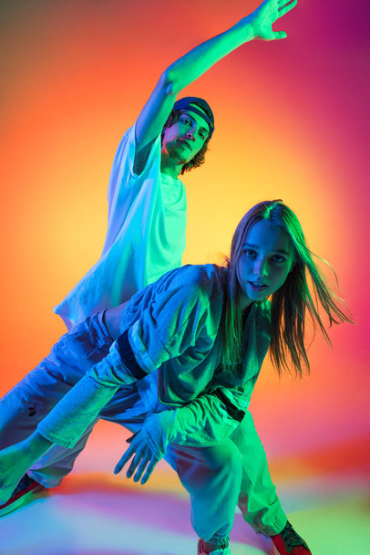 Close-up two hip-hop dancers, stylish emotive girl and boy in action and motion in casual sports youth clothes on gradient multi colored background at dance hall in neon light. - Foto, Imagen