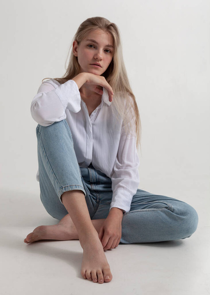 beautiful blonde in a white shirt and jeans sits on the floor - 写真・画像