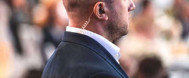 Secret guard service. Private bodyguard with earpiece standing among crowd. Safety of govern and business meeting. Secret service agent listening to his earpiece, side - Photo, Image