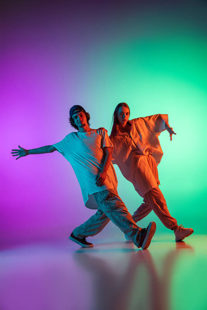 Stylish young hip-hop dancers, emotive girl and boy in action and motion in casual sports youth clothes on gradient multi colored background at dance hall in neon light. - Fotoğraf, Görsel