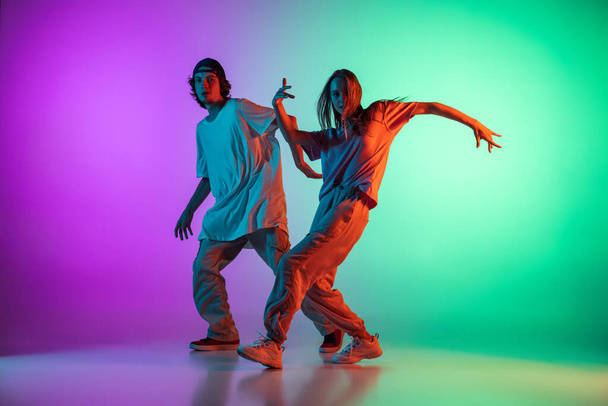 Young hip-hop dancers, stylish emotive girl and boy in action and motion in casual sports youth clothes on gradient multi colored background at dance hall in neon light. - Photo, Image