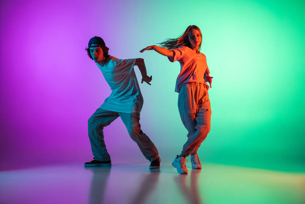 Stylish young hip-hop dancers, emotive girl and boy in action and motion in casual sports youth clothes on gradient multi colored background at dance hall in neon light. - Фото, изображение