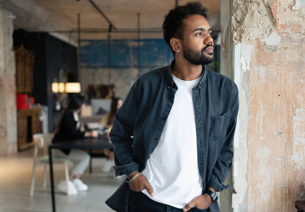 Portrait of mixed-race young man standing in office with colleagues meeting in background - Foto, Bild
