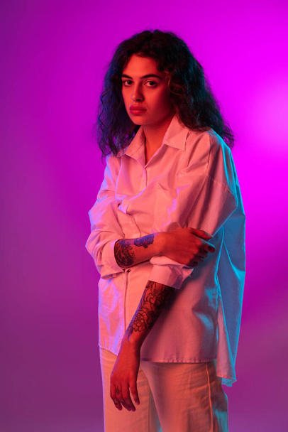 Charming young woman looking at camera isolated on purple, lilac color studio background in neon light. Concept of emotions, expressions - Foto, imagen