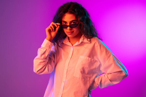 Portrait of beautiful girl putting sunglasses on and looking at camera isolated on purple, lilac color studio background in neon light. - Fotoğraf, Görsel