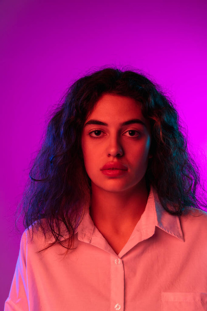 Charming young woman looking at camera isolated on purple, lilac color studio background in neon light. Concept of emotions, expressions - Foto, imagen