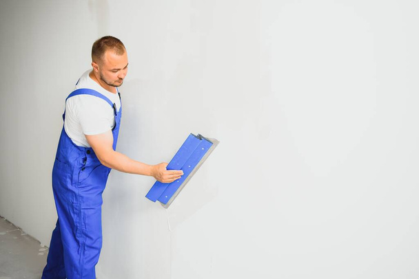 The builder carefully corrects the irregularities of the wall with a trowel. Builder in work clothes against a gray wall. Photo plasterer at work. - Foto, Bild