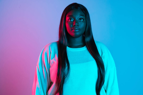 Close-up shot of African charming woman with long straight hair isolated on blue studio background in neon light. Concept of human emotions - Фото, зображення
