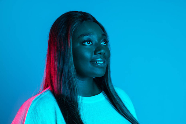 Close-up shot of African charming woman with long straight hair isolated on blue studio background in neon light. Concept of human emotions - Foto, Imagem