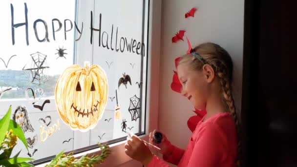 Kind in duivelshoorns die pompoen op het raam schilderen om Halloween te vieren. Little kid tekent decoreert kamer interieur papier vleermuizen viering herfst vakantie huis Creatieve vrije tijd lockdown nieuwe realiteit - Video