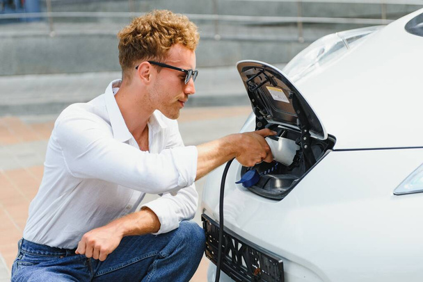 Hansome guy sitting near his new modern electric car and holding plug of the charger, while car is charging at the charging station - Foto, afbeelding