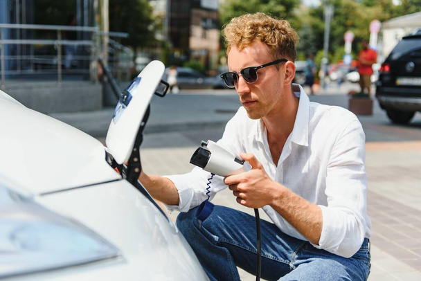 Hansome guy sitting near his new modern electric car and holding plug of the charger, while car is charging at the charging station - Photo, Image