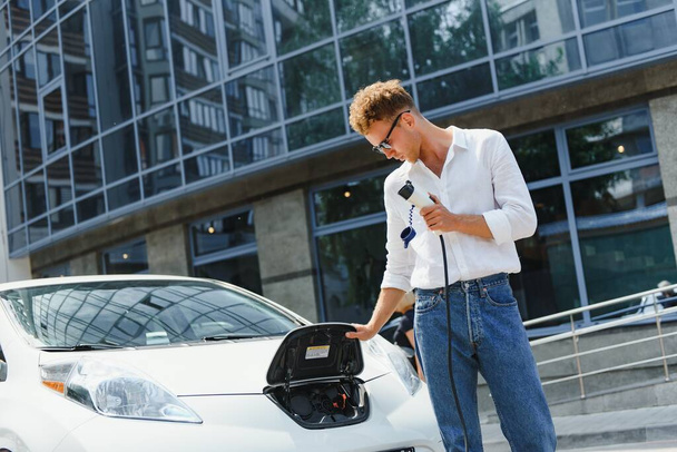 man charging his luxury electric car at outdoor station in front of modern new city buildings - Foto, Imagem