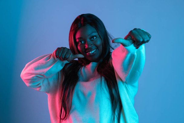 One joyful African beautiful woman with long straight hair singing isolated on blue studio background in neon light. Concept of human emotions - Fotoğraf, Görsel
