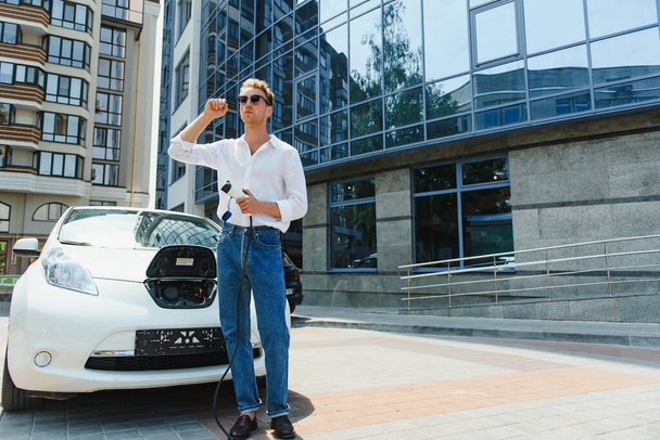Man Holding Power Charging Cable For Electric Car In Outdoor Car Park. And he s going to connect the car to the charging station in the parking lot near the shopping center - Photo, image