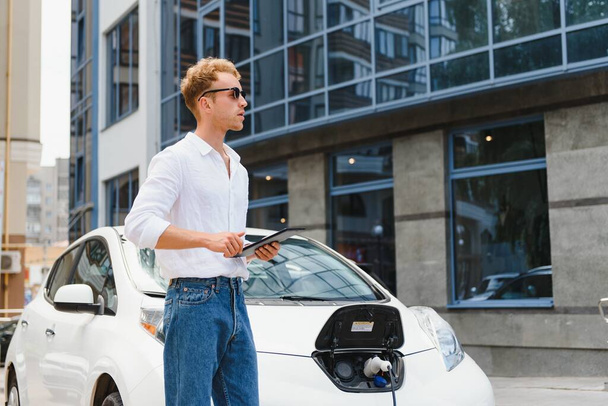 Business man standing near charging electric car and using tablet in the street. - Foto, imagen