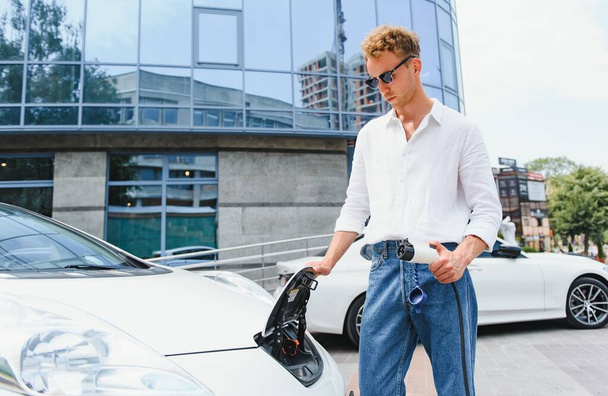 young handsome man holding charging cable at electric charging station point standing near his new car , looking satisfied. - Foto, Imagem