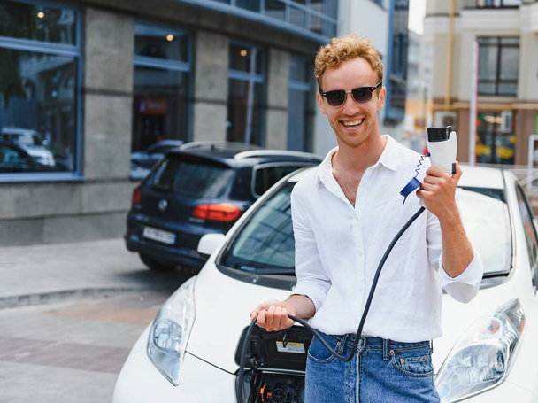 portrait of young handsome man in casual wear, standing at the charging station. Eco electric car concept - Photo, Image