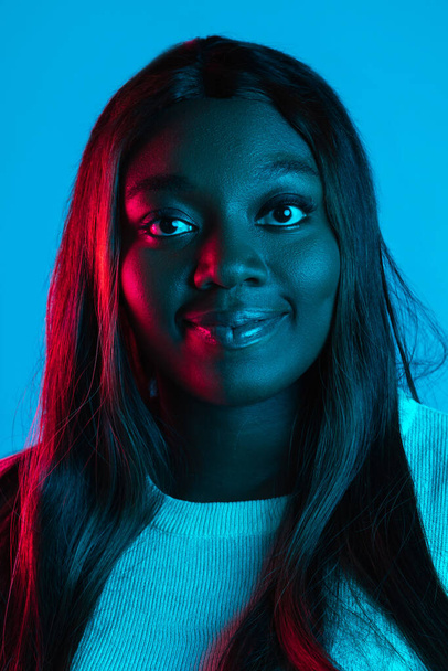 Close-up portrait of African charming woman with long straight hair isolated on blue studio background in neon light. Concept of human emotions - Fotografie, Obrázek