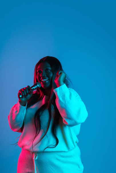 One joyful African beautiful woman with long straight hair singing isolated on blue studio background in neon light. Concept of human emotions - Fotó, kép