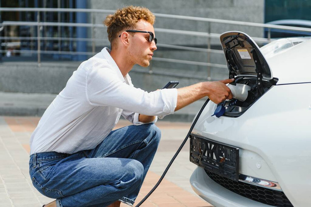 mladý pohledný muž držící nabíjecí kabel na elektrickém nabíjecím stanovišti, stojící poblíž jeho nového auta, vypadá spokojeně. - Fotografie, Obrázek