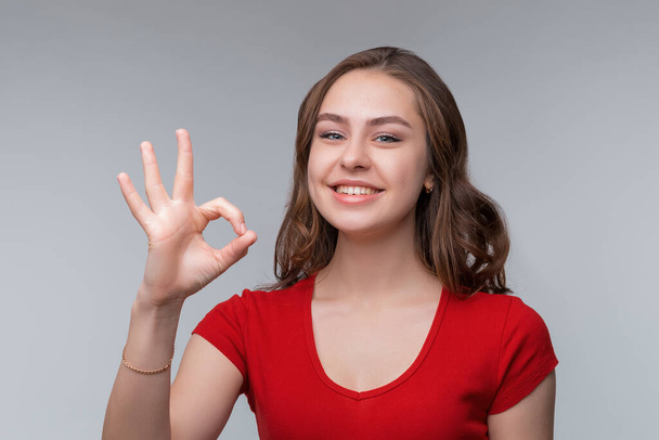 Excellent job. Attractive young brunette woman in red t shirt showing OK sign and smiling satisfied, give positive reply, show alright agree sign, making okay and nod in approval, standing against gray background - Foto, Imagen