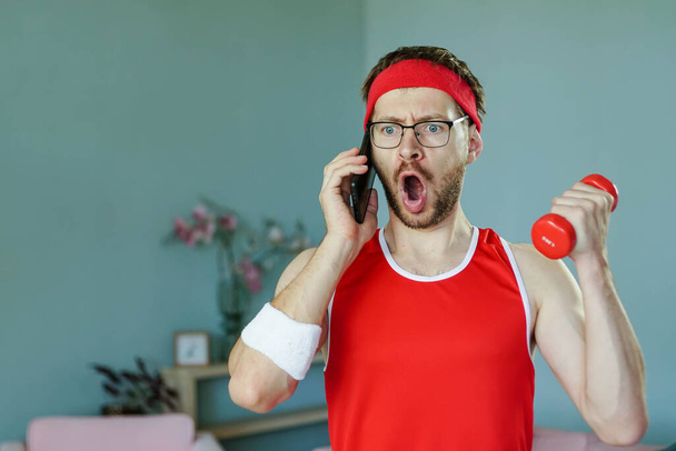 Portrait of funny surprised geek talking on the phone while exercising with dumbbell - Фото, зображення