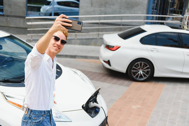 Portrait of young handsome european man, making selfie photo on his smartphone while leaning on his electric car, charging the battery at city power station after buying frsh products in trade mall - Foto, Imagem